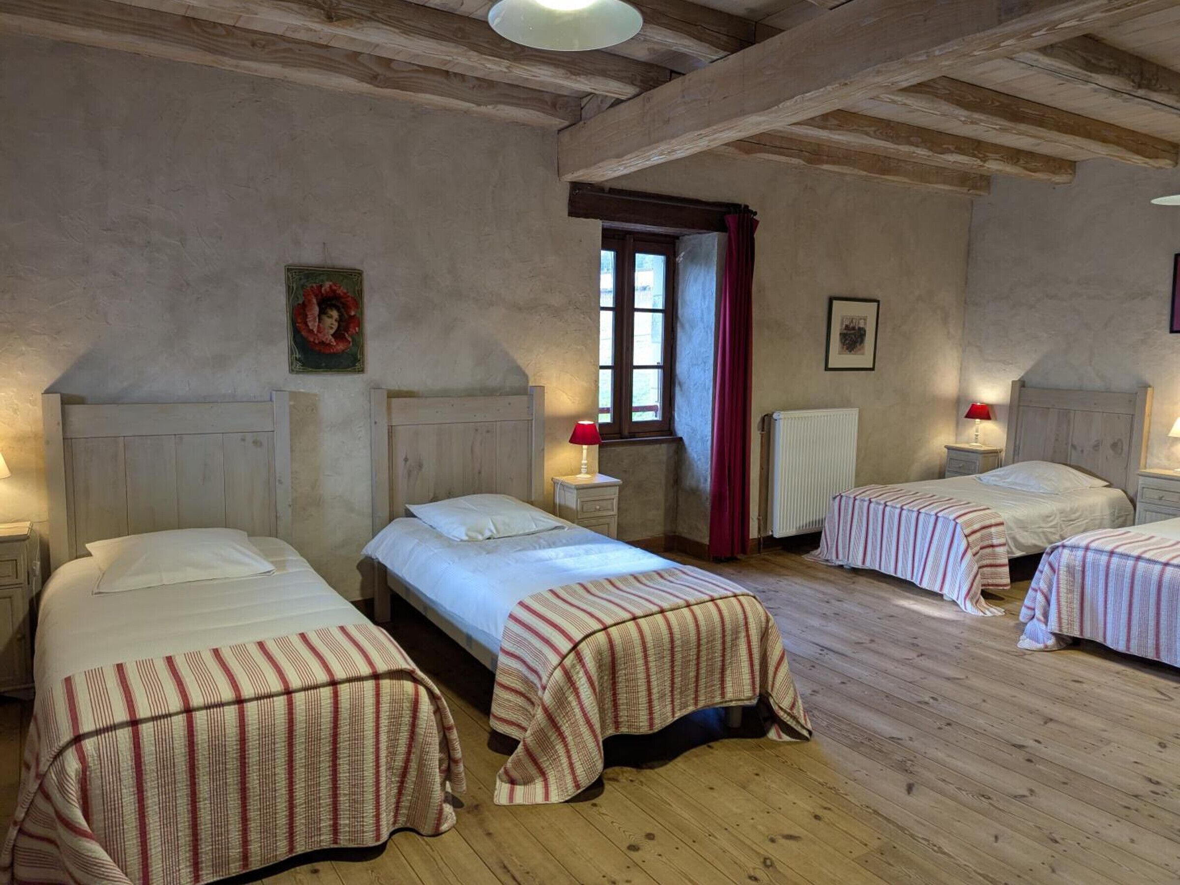 Une chambre avec trois lits, des poutres en bois au plafond, une fenêtre avec des rideaux rouges et un radiateur.
