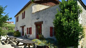 A rustic stone building with a sloped roof, red shutters, and a small garden area with a barbecue.