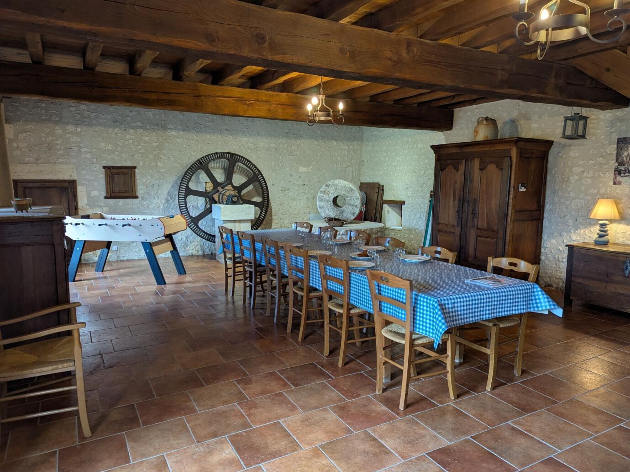 Un comedor rústico con una gran mesa de madera dispuesta para seis personas, rodeada de sillas. Se ve un aparador de madera y un reloj colgado en la pared.