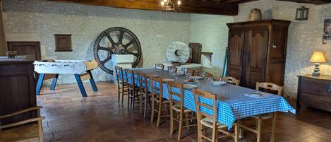 Un comedor rústico con una gran mesa de madera dispuesta para seis personas, rodeada de sillas. Se ve un aparador de madera y un reloj colgado en la pared.