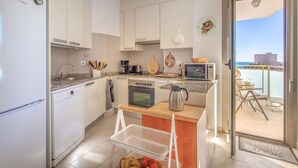 A modern kitchen with white cabinets, a stove, microwave, and a coffee maker. There's a small dining area with a table and chairs, and a view of the sea through the glass door. - Cozy apartment in El Campello with WiFi (El Campello)