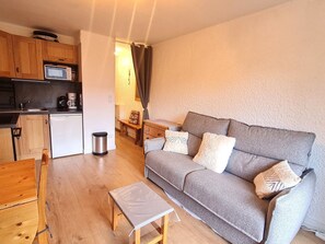 A compact living space with a kitchenette, a grey sofa with patterned cushions, a small wooden table, and a bench.