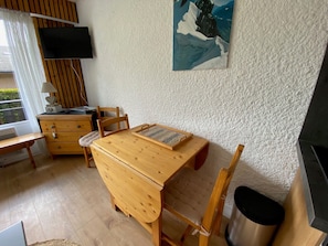 Una habitación acogedora con una mesa de madera, una silla, un mueble pequeño, un televisor montado en la pared y un cuadro sobre la mesa.