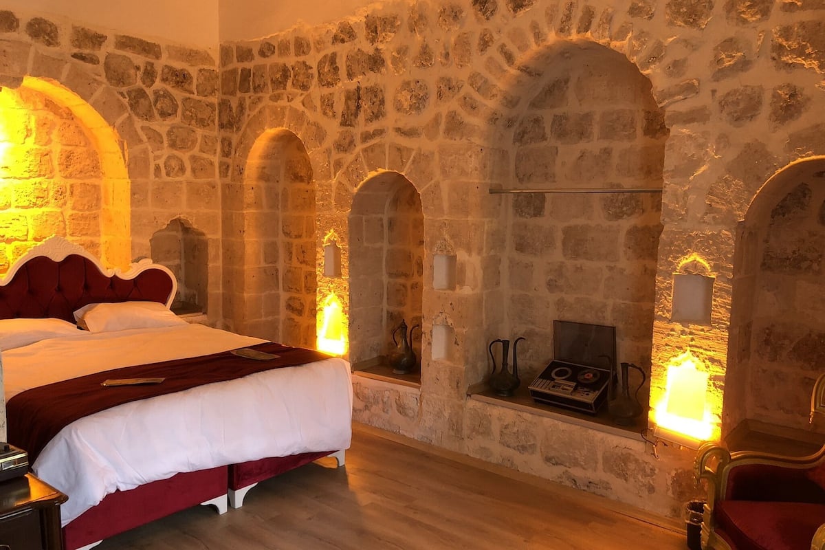 A room with a stone wall, a bed with a red headboard, a fireplace, and a red armchair.