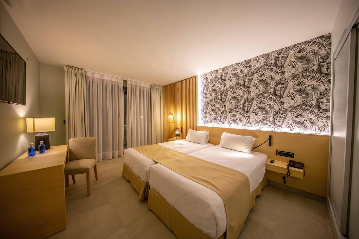 Une chambre d’hôtel avec un grand lit, un bureau, une chaise, une table de chevet et un mur recouvert d’un papier peint aux motifs tropicaux.
