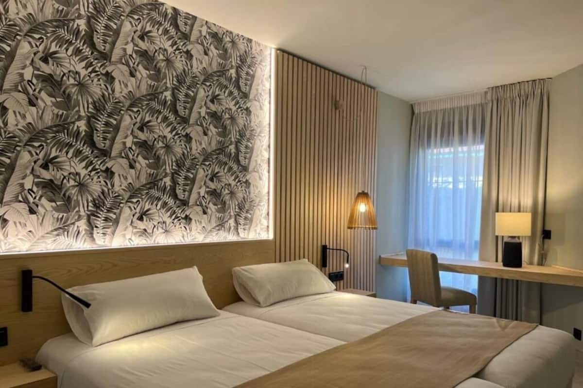 Une chambre d’hôtel comprenant un lit, un bureau, une chaise, une lampe et un mur orné d’un motif de feuilles.
