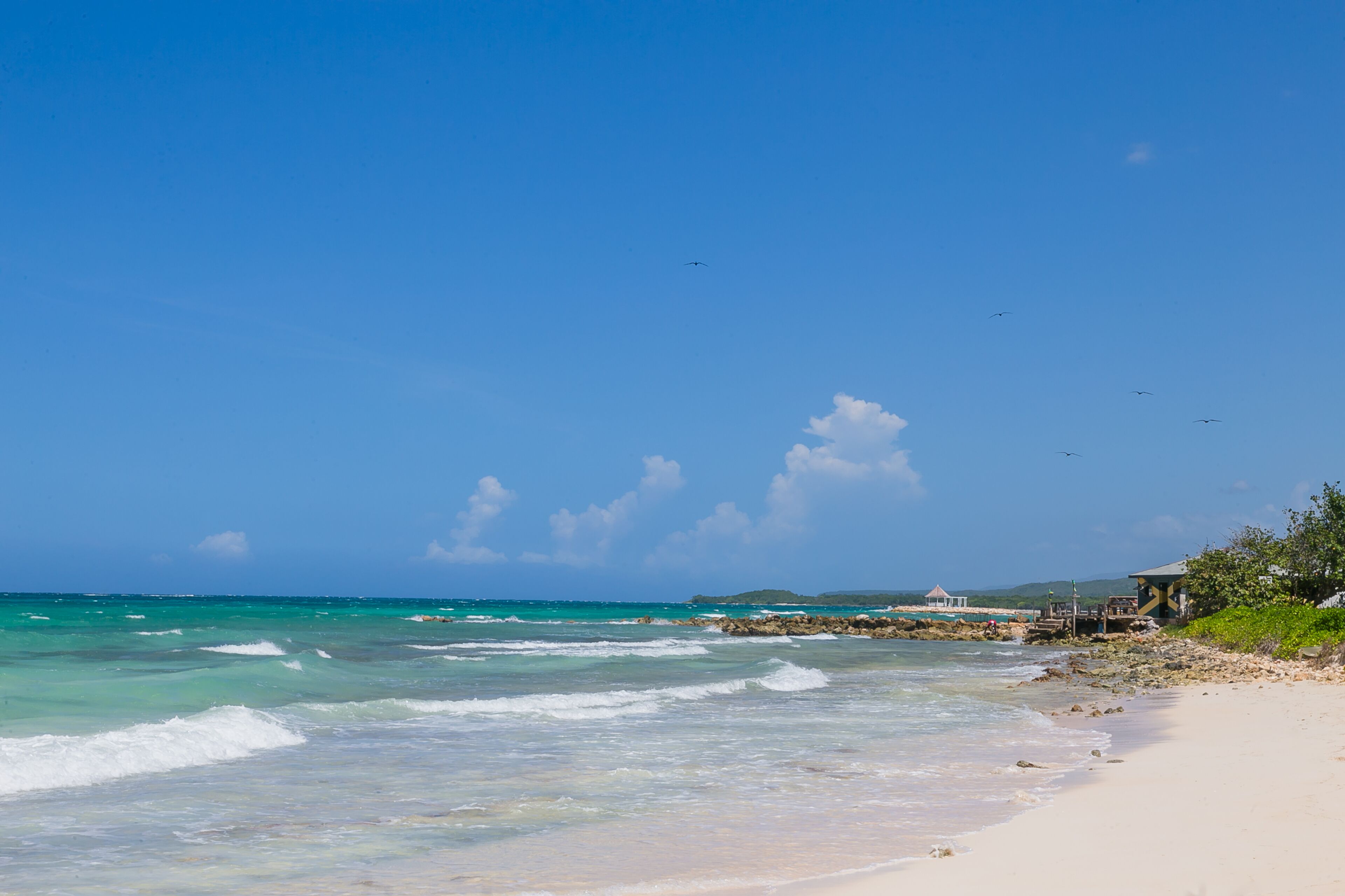 Ein Strand mit klarem türkisblauem Wasser, einem sandigen Ufer und einer kleinen, überdachten Struktur.