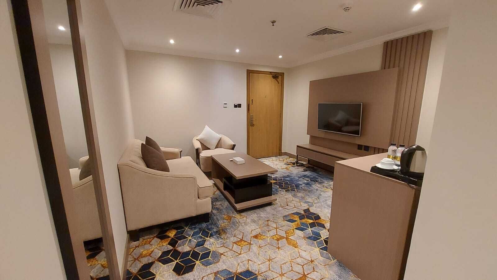A modern hotel room with a sofa, armchair, coffee table, TV, and a minibar.
