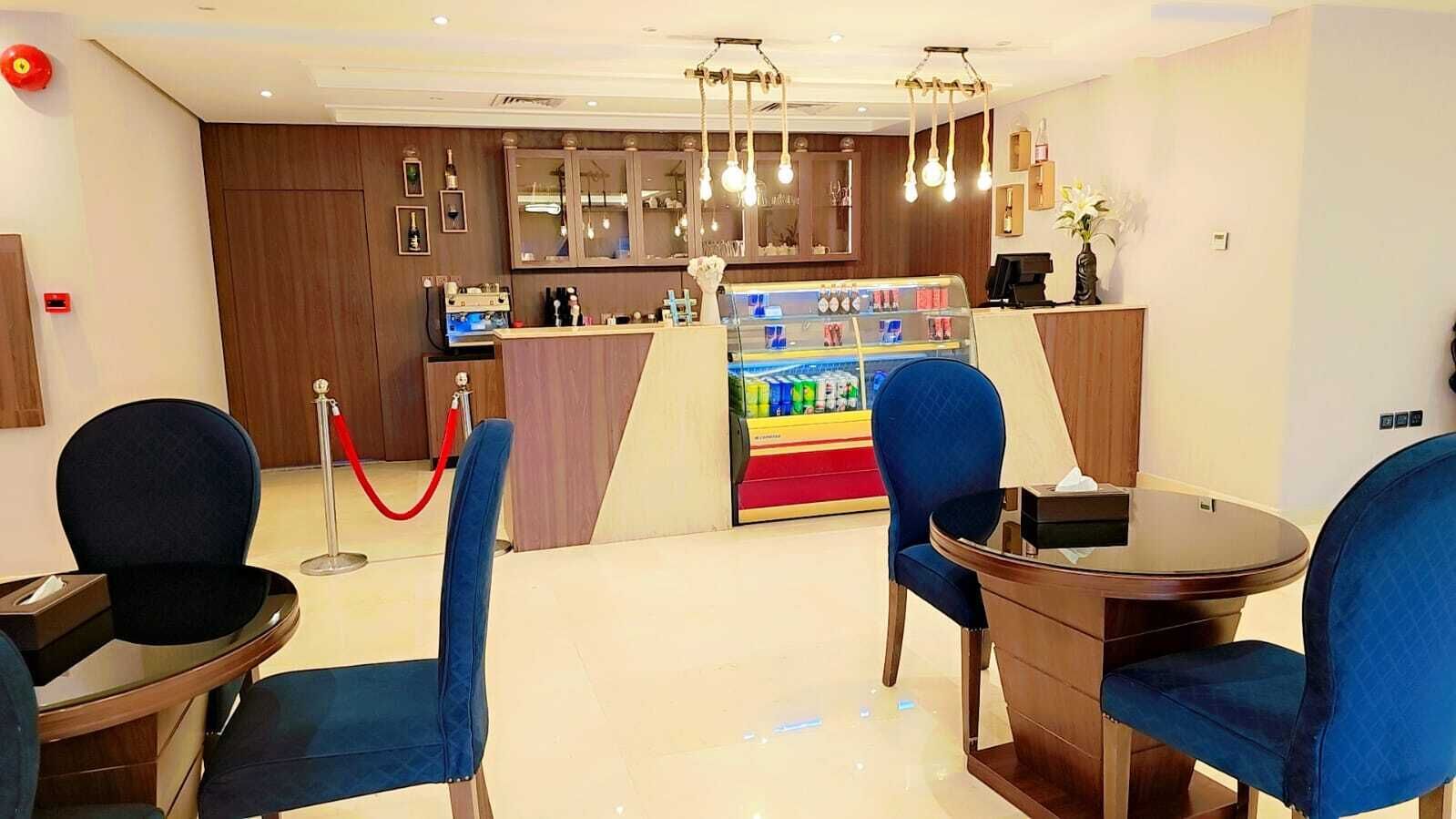 A hotel lobby with a reception desk, blue upholstered chairs, a small bar area, and a red rope barrier.