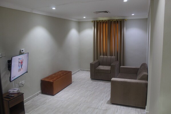 A modern living room with a flat-screen TV mounted on the wall, a brown ottoman, and two beige armchairs. - ماجيك سويت الرقعى Magic Suite ALRaggi (Kuwait City)