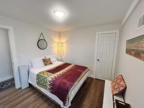Un dormitorio con cama, mesita de noche, lámpara colgante, puerta y una silla pequeña.