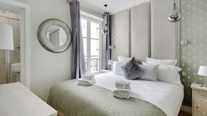 A bedroom with a bed, a round mirror, a window with curtains, a bedside table, and a hanging light fixture. - Rue Lepic - quintessential winding cobblestone street- Take a step into the past (Paris)