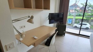 A hotel room with a wooden desk, a chair, a TV, and a large window with a view of the outside. - Naiika Co Living (Denpasar)