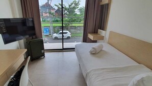 A hotel room with a bed, a desk with a TV, a suitcase, and a view of a street and greenery outside. - Naiika Co Living (Denpasar)