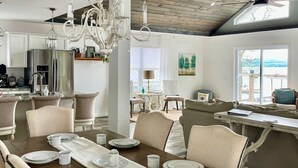 Comedor con mesa de madera dispuesta para cuatro personas, sillas tapizadas, una araña y vistas al exterior a través de amplios ventanales.