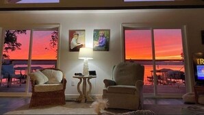 Una sala de estar con dos sillones, una mesa y una lámpara. Se ven dos ventanas con vistas al atardecer.