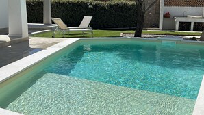 Une piscine rectangulaire à l’eau claire, entourée d’une terrasse carrelée de blanc et agrémentée de deux chaises longues blanches.