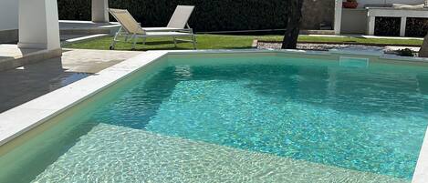 A rectangular swimming pool with clear water, surrounded by a white tiled deck and two white lounge chairs.