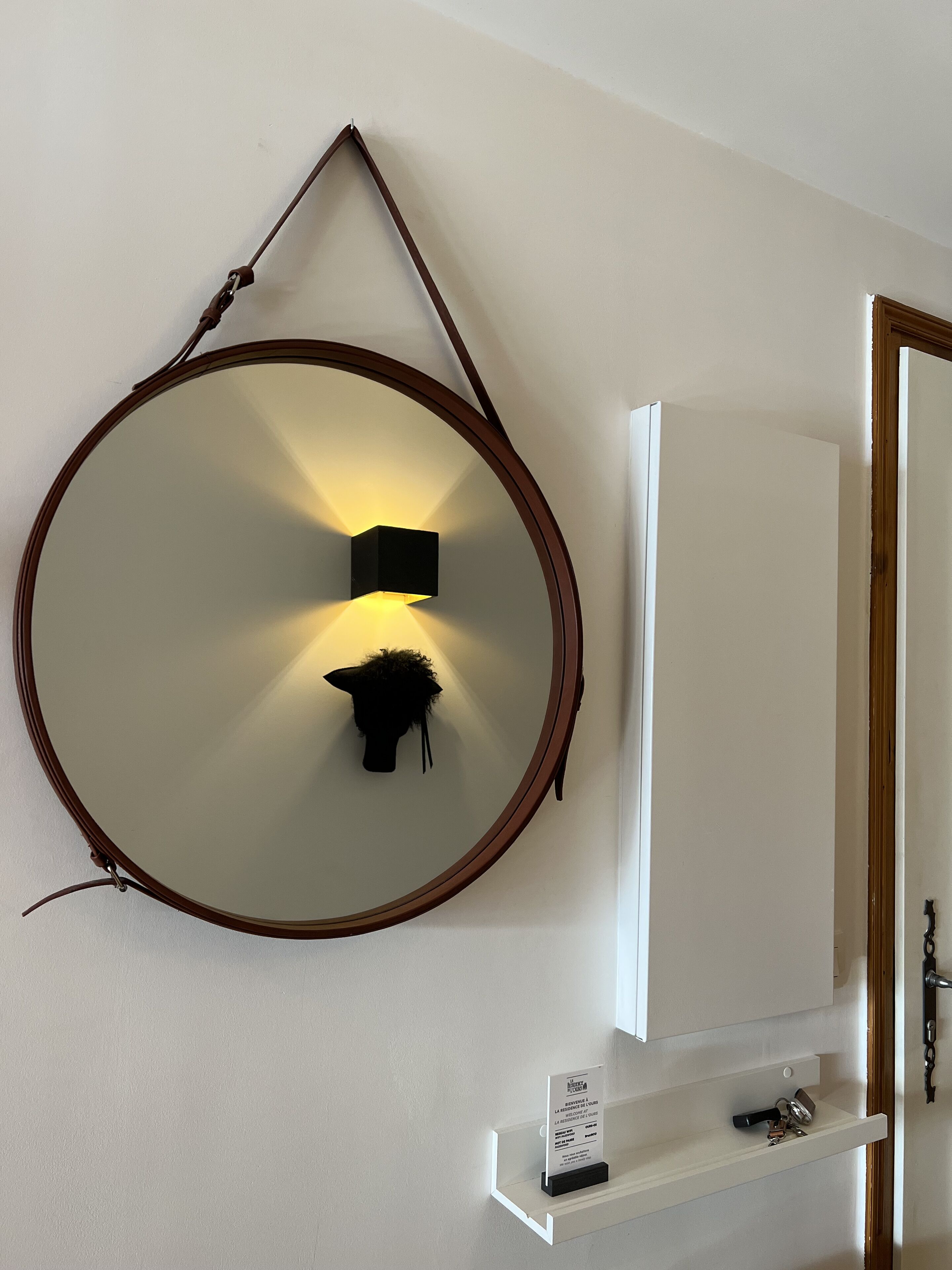 A round mirror with a brown frame, a wall-mounted light, and a small shelf below.