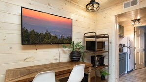 Eine gemütliche Küche mit einem Holztisch, zwei weißen Stühlen, einem an der Wand befestigten Fernseher und einer Pflanze auf dem Tisch.