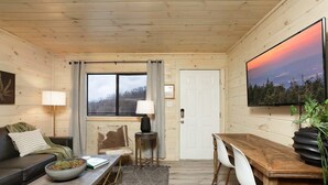 Ein gemütliches Blockhaus-Interieur mit Holzboden, einer Couch, einem Esstisch mit Stühlen, einem an der Wand befestigten Fernseher und einem Fenster mit Vorhängen.