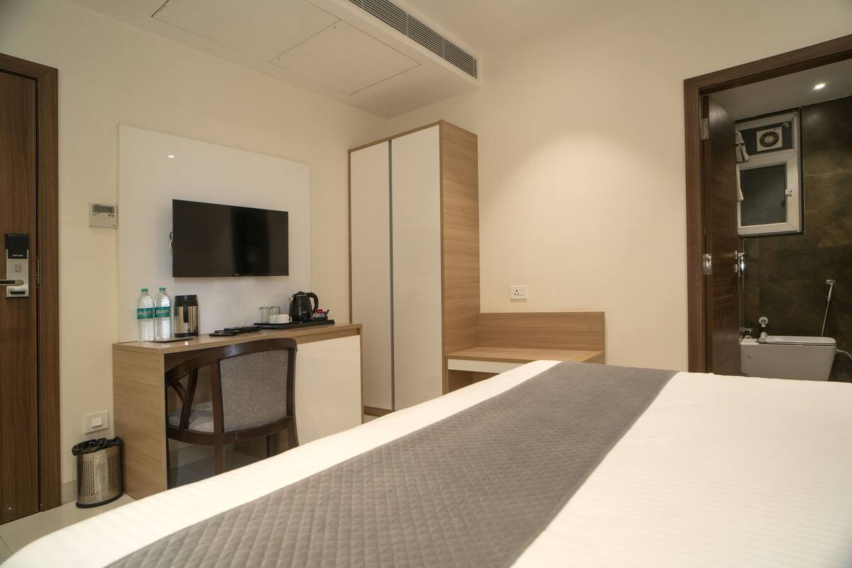 A hotel room with a bed, a desk with a chair, a television, a wardrobe, and a bathroom visible through an open door.