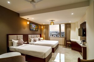 Quarto de hotel com duas camas, uma escrivaninha, uma cadeira, uma televisão e uma janela grande com vista para a cidade.
