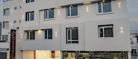 Un hotel de varios pisos con un diseño moderno, que presenta un letrero grande que dice 'HOTEL', una escalera que conduce a la entrada y un puesto de comida visible a nivel de la calle.