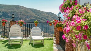 Eine Terrasse mit zwei weißen Stühlen, einer Steinmauer und hängenden Blumenampeln.