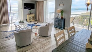 Ett rymligt vardagsrum med en stor TV, en matplats och en balkong med utsikt över stranden.