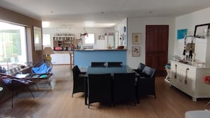 A spacious living room with a dining area, a kitchen in the background, and a large window providing natural light. - Villa 130 m2, pool and terrace, facing the sea, on 1300m2 of wooded grounds. (Hyères)