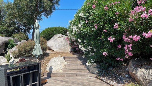 A wooden deck leading to a seating area with a wicker chair and a table, surrounded by rocks and flowering plants. - Villa Avec Piscine (Porto-Vecchio)