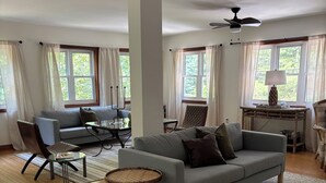 A spacious living room with a grey sofa, a small round table, and a standing lamp. There are two windows with curtains, a ceiling fan, and a patterned rug on the wooden floor.