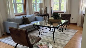 A living room with a sofa, two armchairs, a glass coffee table, and a rug. There are windows with curtains, a lamp, and a small plant on a side table.