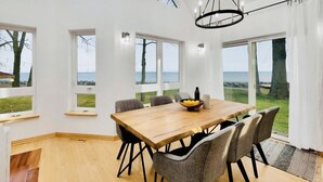 Ein Essbereich mit einem Holztisch, grauen Stühlen und einem Kronleuchter. Große Fenster bieten Blick auf das Meer.