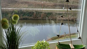 Vista desde la ventana: un lago con colinas al fondo, un jarrón verde con flores y una pequeña decoración de un velero.