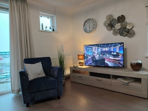 Ein Wohnzimmer mit blauem Sessel, Fernseher, der eine Nachrichtensendung zeigt, Wanduhr, dekorativen Tellern und einem Fenster mit Blick nach draußen.