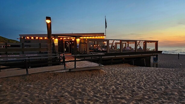 Un restaurante frente a la playa con mesas al aire libre, vistas al océano, una terraza de madera y una playa arenosa en primer plano.