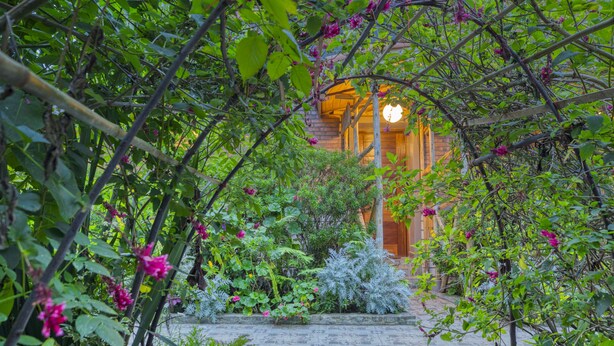 Un chemin de pierres mène à un bâtiment en bois dont l’intérieur est éclairé d’une lumière chaleureuse, visible à travers une arche.