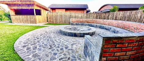 A circular stone fire pit with a brick wall surrounding it, set in a grassy backyard with wooden fences and buildings in the background.