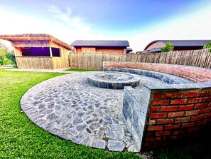 A circular stone fire pit with a brick wall surrounding it, set in a grassy backyard with wooden fences and buildings in the background. - Virunga Inn Resort & SPA (Musanze)