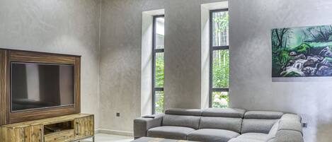 A modern living room with a sectional sofa, a flat-screen TV on a wooden cabinet, and a painting of a forest scene.