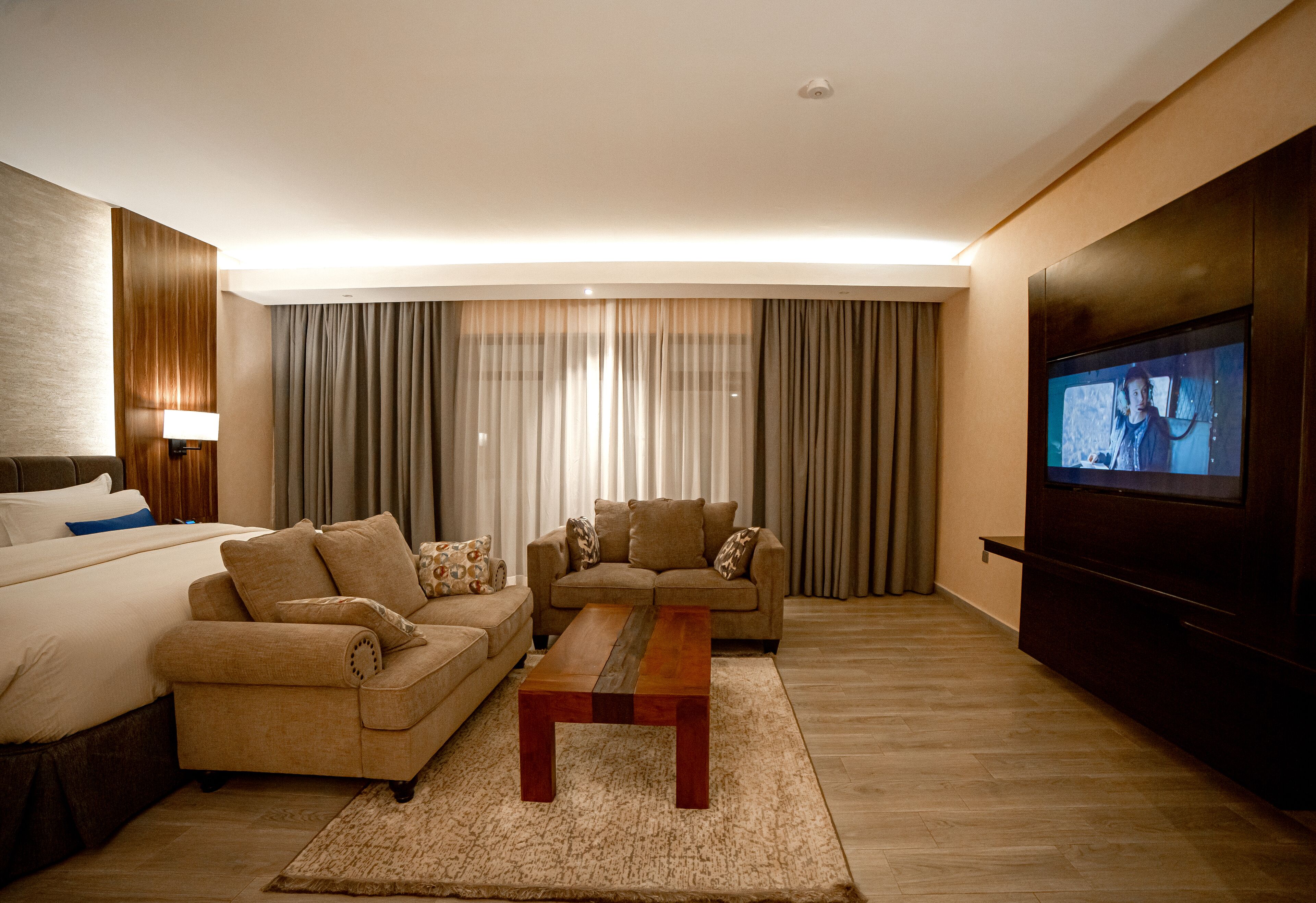 A hotel room with a bed, two sofas, a coffee table, a TV, and a large window with curtains.