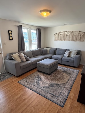 A modern living room with a grey sectional sofa, a matching ottoman, a patterned rug, a macrame wall hanging, and a window with curtains.
