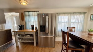 Una cocina compacta con un refrigerador de acero inoxidable, una cafetera y un dispensador de agua. Hay una mesa de madera con sillas y una pequeña estantería debajo.