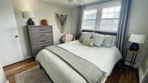 Un dormitorio con cama, cómoda, silla, ventilador de techo y ventana con cortinas.