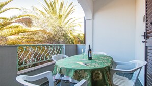 Une petite terrasse extérieure avec une table ronde recouverte d’une nappe verte, des chaises en plastique blanches et une bouteille de vin.
