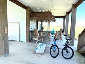 A covered outdoor area with a wooden table and chairs, a thatched roof, and a bicycle. - *Brand New* Luxury Beach Cottage in Port Bolivar! Short Walk to the Beach! (Bolivar Peninsula)