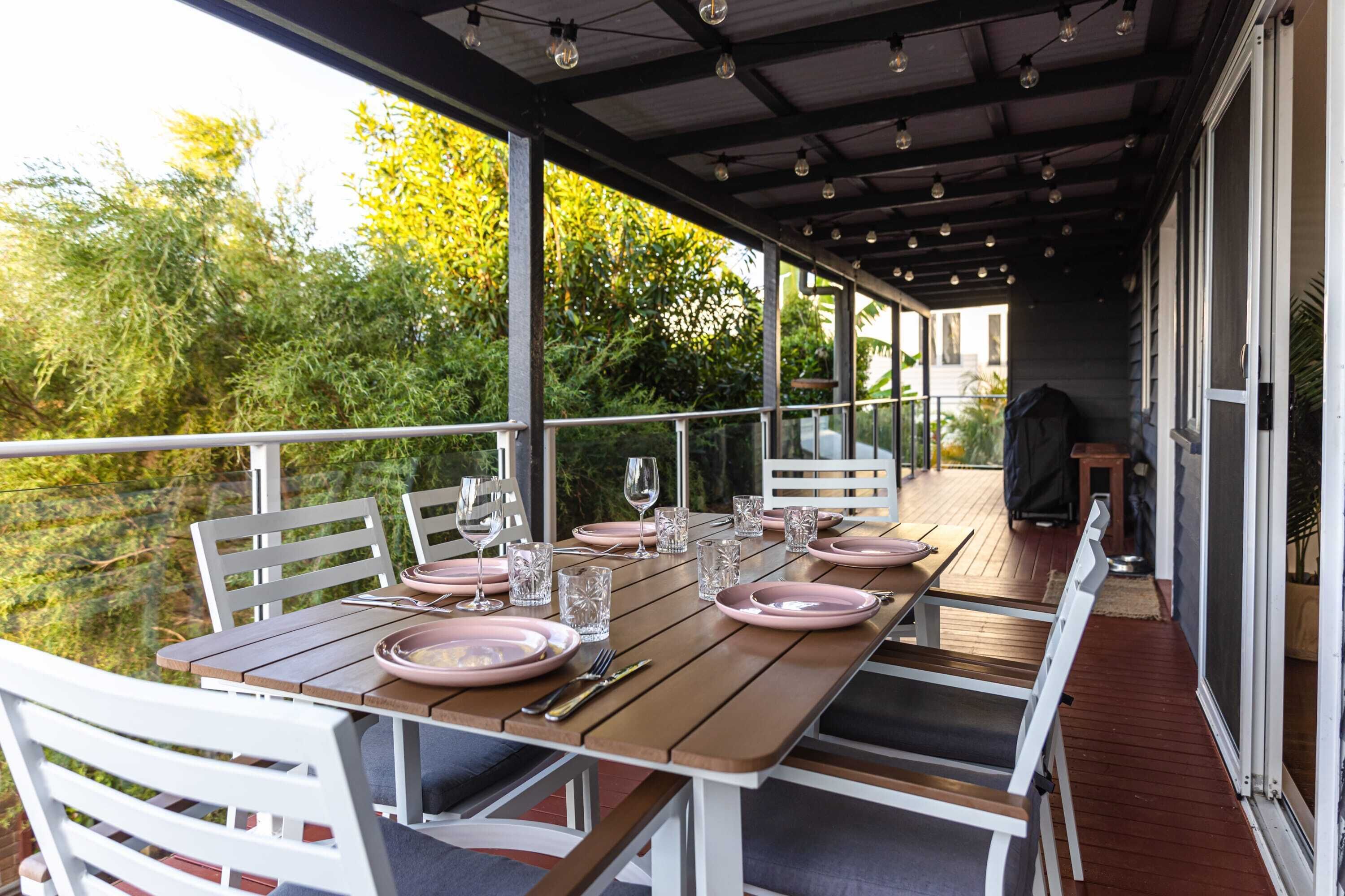 Un espace extérieur abrité, aménagé pour les repas, avec une table en bois prévue pour quatre personnes, entourée de chaises blanches, et donnant sur un jardin luxuriant.