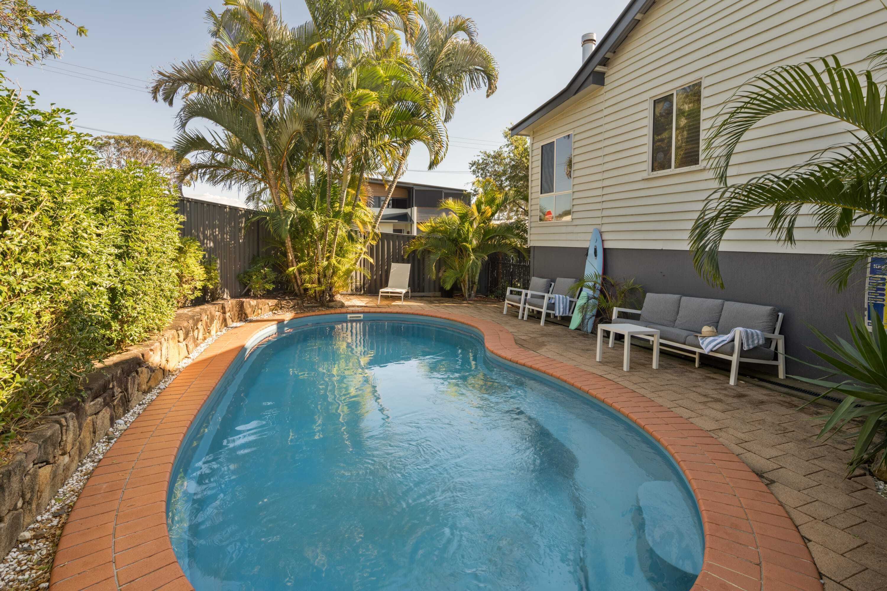 A swimming pool with a brick border, surrounded by a patio area with a sofa and chairs.
